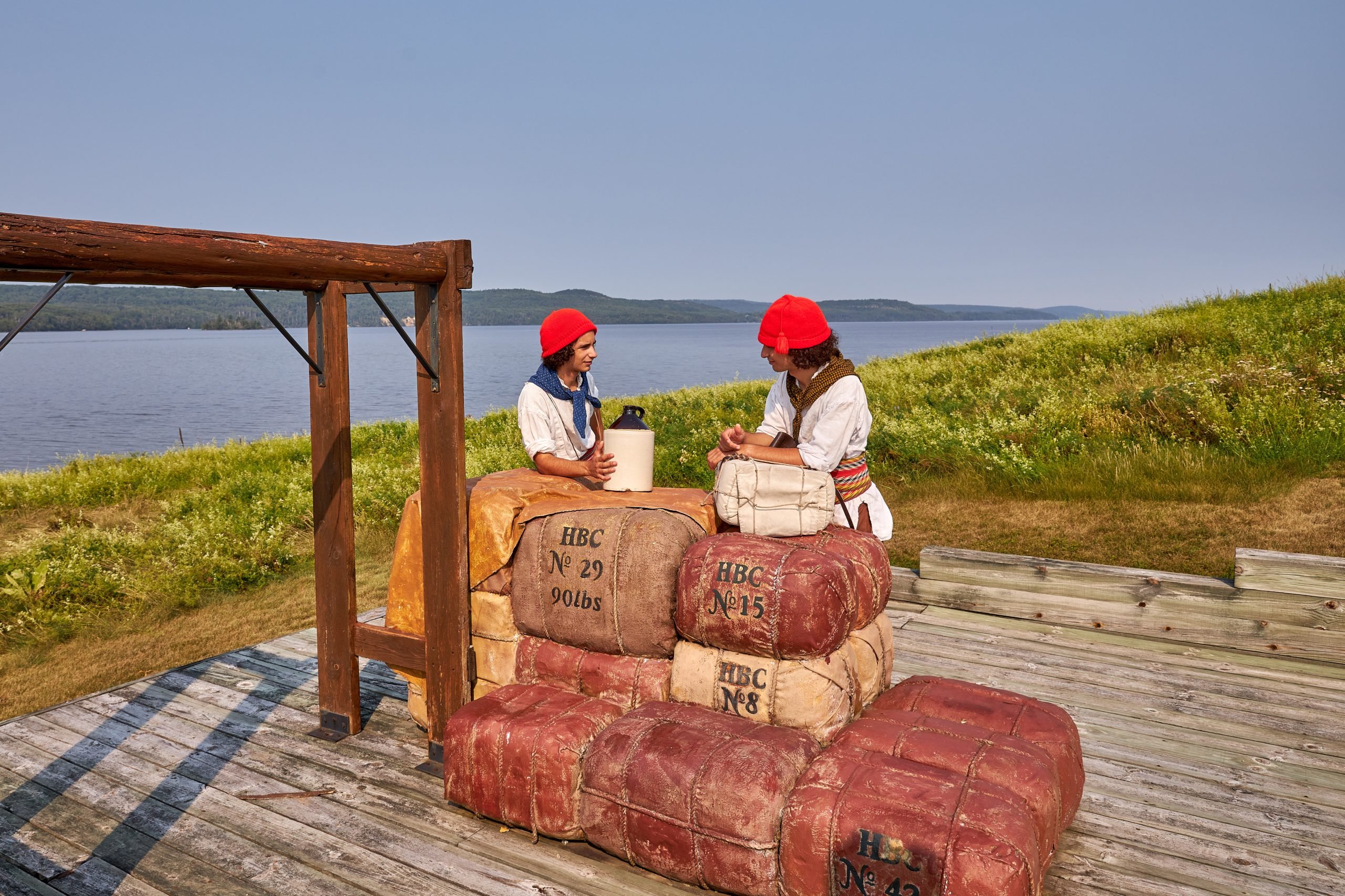 Deux statuts représentant les premiers arrivants blanc sur les rives du la Témiscamingue. Ceux-ci semblent marchander des biens qui se trouvent sous forment de ballot. 
